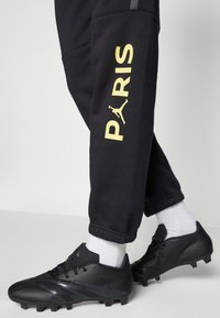 Czarne spodnie sportowe z napisem „PARIS” i żółtym logo, zestawione z czarnymi korkami piłkarskimi. Posiadają białe skarpety i elastyczne mankiety przy nogawkach.