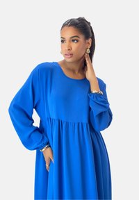 Lange blauwe jurk met een ronde hals, gerimpelde taille en ballonmouwen. Soepele stof met een lichte textuur. Zilveren accessoires zichtbaar.