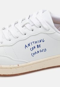 White leather sneaker with round toe, flat white laces, and blue text "ANYTHING CAN BE CHANGED" on the side. Textured rubber sole.