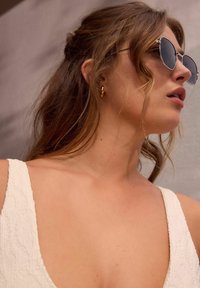 Gafas de sol redondas con lentes oscuros, montura de metal plateado y una prenda blanca texturizada. Se ve piel suave y cabello castaño ondulado.