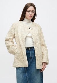 Vrouw die een roomkleurige, textuurrijke wollen jas draagt over een witte coltrui en een high-waisted spijkerrok met een zwarte riem, staand tegen een witte achtergrond.