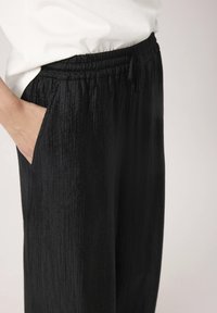 Schwarze, strukturierte Hose mit lockerem Schnitt, die einen elastischen Bund und Seitentaschen hat. Der Stoff hat eine leichte, zerknitterte Optik.
