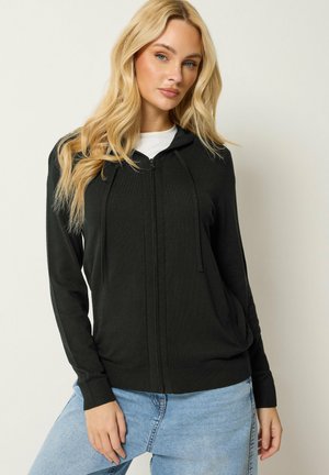 Blonde Frau, die einen schwarzen Kapuzenpullover mit Reißverschluss über einem weißen Shirt und hellblauen Jeans trägt, vor einem schlichten hellen Hintergrund stehend.
