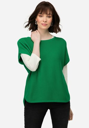 Grünes, kurzärmliges Oberteil mit gerippter Textur, das über einem weißen Langarmshirt getragen wird. Hat einen runden Ausschnitt und dezente seitliche Schlitze.