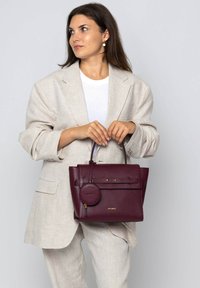 Sac à main en cuir bordeaux avec un design structuré, un pendentif rond amovible et des accents dorés. Associé à un blazer beige et un haut blanc.
