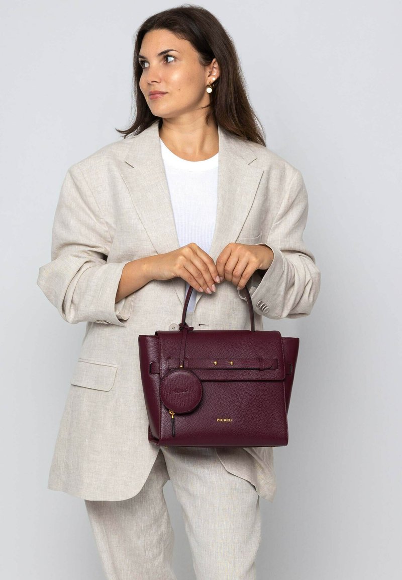 Sac à main en cuir bordeaux avec un design structuré, un pendentif rond amovible et des accents dorés. Associé à un blazer beige et un haut blanc.