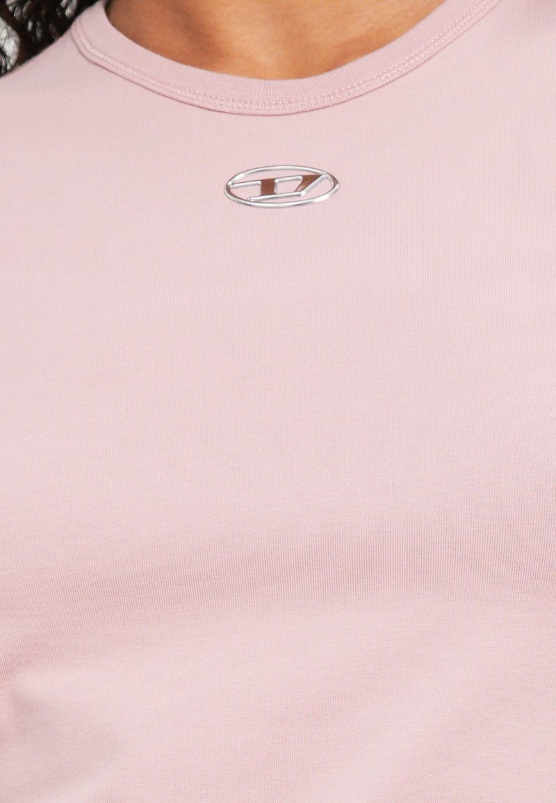 Nærbillede af en person iført en lyserød skjorte med en lille sølvfarvet oval logo med et stiliseret "H" nær halsudskæringen.