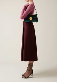 Sac en daim vert avec un fermoir circulaire doré, porté avec une longue jupe bordeaux et un haut en tricot rose clair. Sandales à talons brunes et chaussettes.