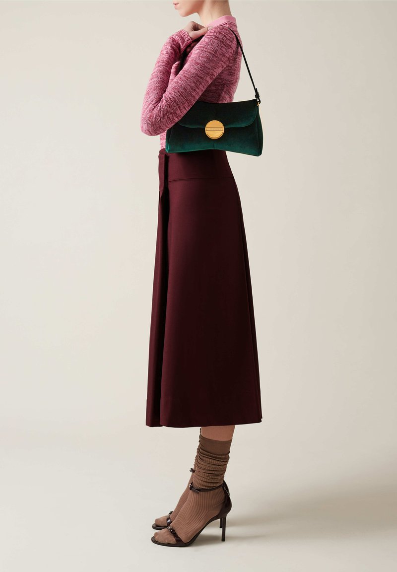 Sac en daim vert avec un fermoir circulaire doré, porté avec une longue jupe bordeaux et un haut en tricot rose clair. Sandales à talons brunes et chaussettes.