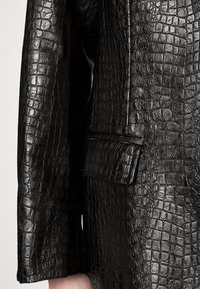 Veste en cuir noir avec un motif texturé de crocodile, dotée d'une poche à rabat unique et de manches larges. Finition lisse et brillante.