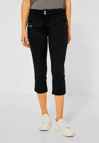 Pantaloni neri a vita alta in tessuto elasticizzato, con tasca con zip e due bottoni in vita. Indossati con sneakers bianche.