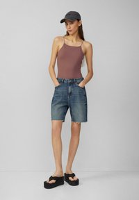 Brązowy top na ramiączkach, szorty dżinsowe z wysokim stanem i postrzępionym dołem oraz czarne sandały na platformie. Model nosi szary kapelusz, pozując na jednolitym tle.