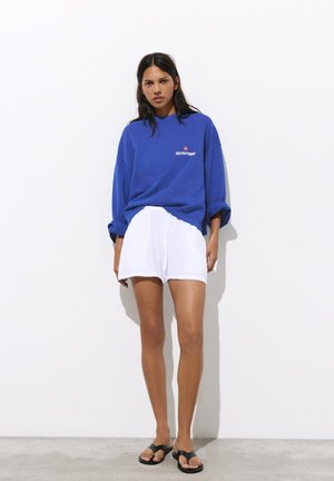 Kvinde, der står mod en hvid væg iført en blå sweatshirt med teksten "Go Outside", hvide shorts og sorte sandaler.