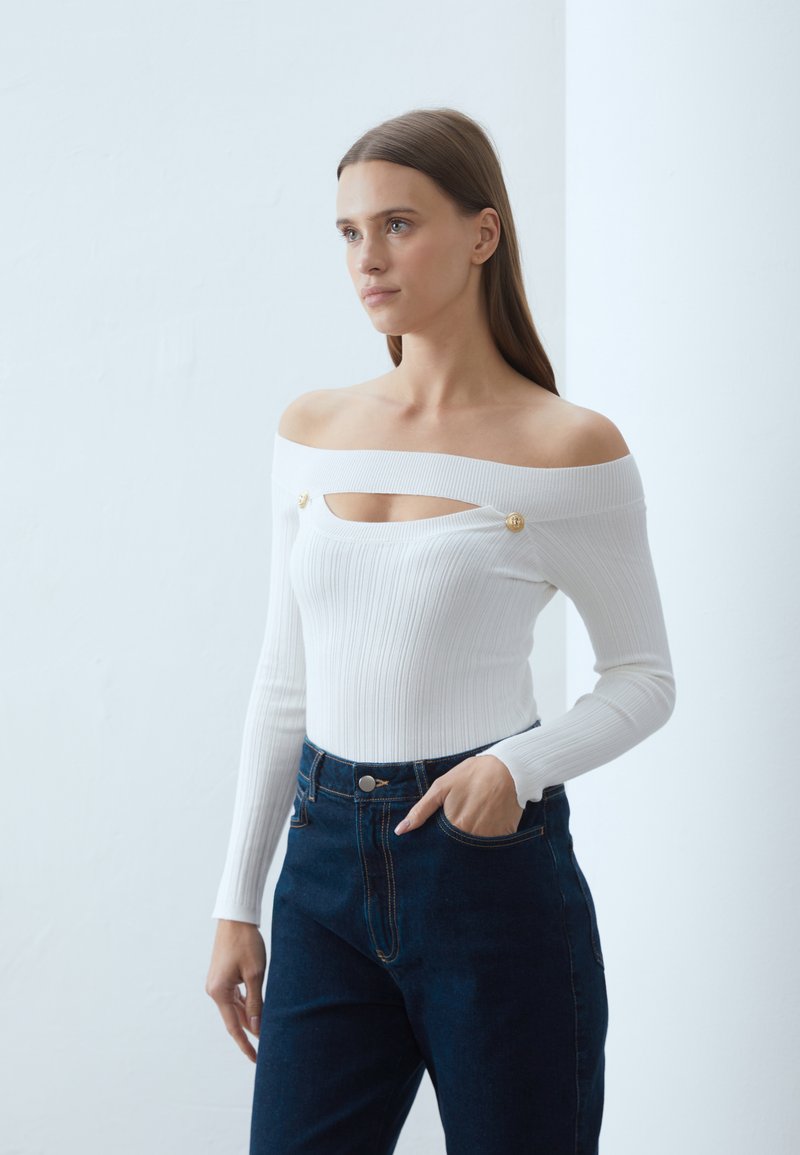 Balts ribots topi bez pleciem ar izgriezumu un zeltītām pogām, kopā ar augstajiem tumšo džinsu bikšu. Gluda tekstūra.