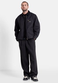 Giacca nera con una tasca con zip, camicia nera sotto, pantaloni neri e scarpe nere lucide. Presenta una vestibilità comoda e un tessuto resistente.