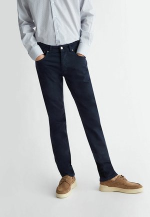 Osoba ubrana w ciemnoniebieskie dopasowane spodnie, jasną wzorzystą koszulę na guziki oraz beżowe zamszowe buty casual, stojąca z rękami w kieszeniach.