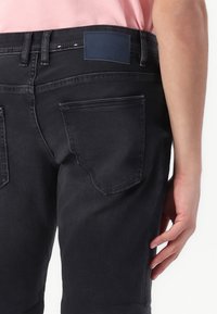 Jeans en denim noir avec deux poches arrière, une étiquette bleu foncé à la taille et une texture lisse. La coupe est ajustée.