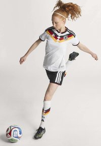 Maglia da calcio femminile: maglietta bianca con un motivo geometrico rosso, giallo e nero, pantaloni corti neri e calze a righe; abbinata a scarpe da calcio nere.