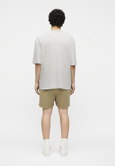 Άτομο με σγουρά μαλλιά που φορά γκρι και λευκό ριγωτό κοντομάνικο μπλουζάκι, χακί σορτς, λευκές κάλτσες και λευκά αθλητικά, με την πλάτη γυρισμένη.