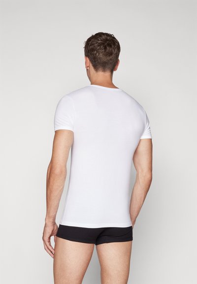 Bela kratka majica s kratkimi rokavi, izdelana iz mehkega bombaža, s okroglim izrezom. Model nosi črne kratke hlače, ki so vidne pri pasu.