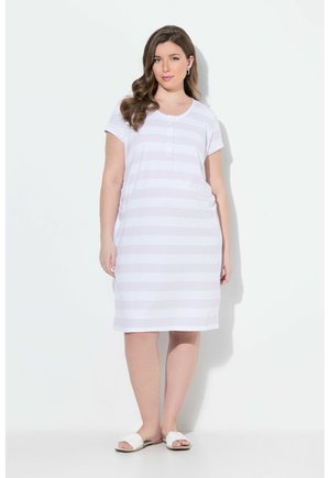 Femme aux cheveux longs et ondulés portant une robe blanche rayée à manches courtes, longueur genou, et des sandales blanches sans lacets, debout devant un fond uni.