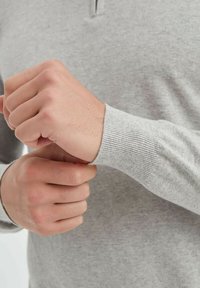 Pull en tricot gris clair avec de longues manches et des poignets à côtes, comportant une fermeture éclair partielle au niveau du col. Gros plan sur des mains ajustant les manches.