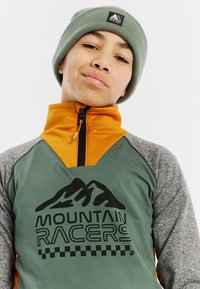 Zelená a oranžová zipsová fleecová bunda so sivými rukávmi, obsahujúca text "MOUNTAIN RACERS" a grafiku hory. Nosí sa so zeleným pleteným čiapkou.