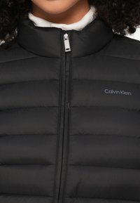 Μαύρο αμάνικο μπουφάν τύπου puffer με ψηλό γιακά, υφή με καπιτονέ ραφές και κλείσιμο με φερμουάρ. Διαθέτει διακριτικό λογότυπο "Calvin Klein" σε γκρι κοντά στο φερμουάρ.