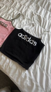Két Adidas ujjatlan póló, az egyik fekete fehér díszítéssel, a másik rózsaszín szürke díszítéssel, laposan fekszik egy fehér texturált takarón.