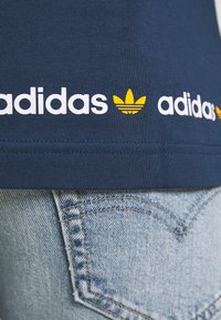 Námořnická modrá bavlněná tričko s bílým a žlutým logem "adidas" a emblémem pod lemem, spárované s světlými modrými džíny.