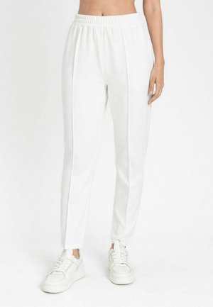 Donna che indossa pantaloni bianchi a vita alta con gamba dritta e cuciture laterali, insieme a sneaker bianche, in piedi davanti a uno sfondo bianco.