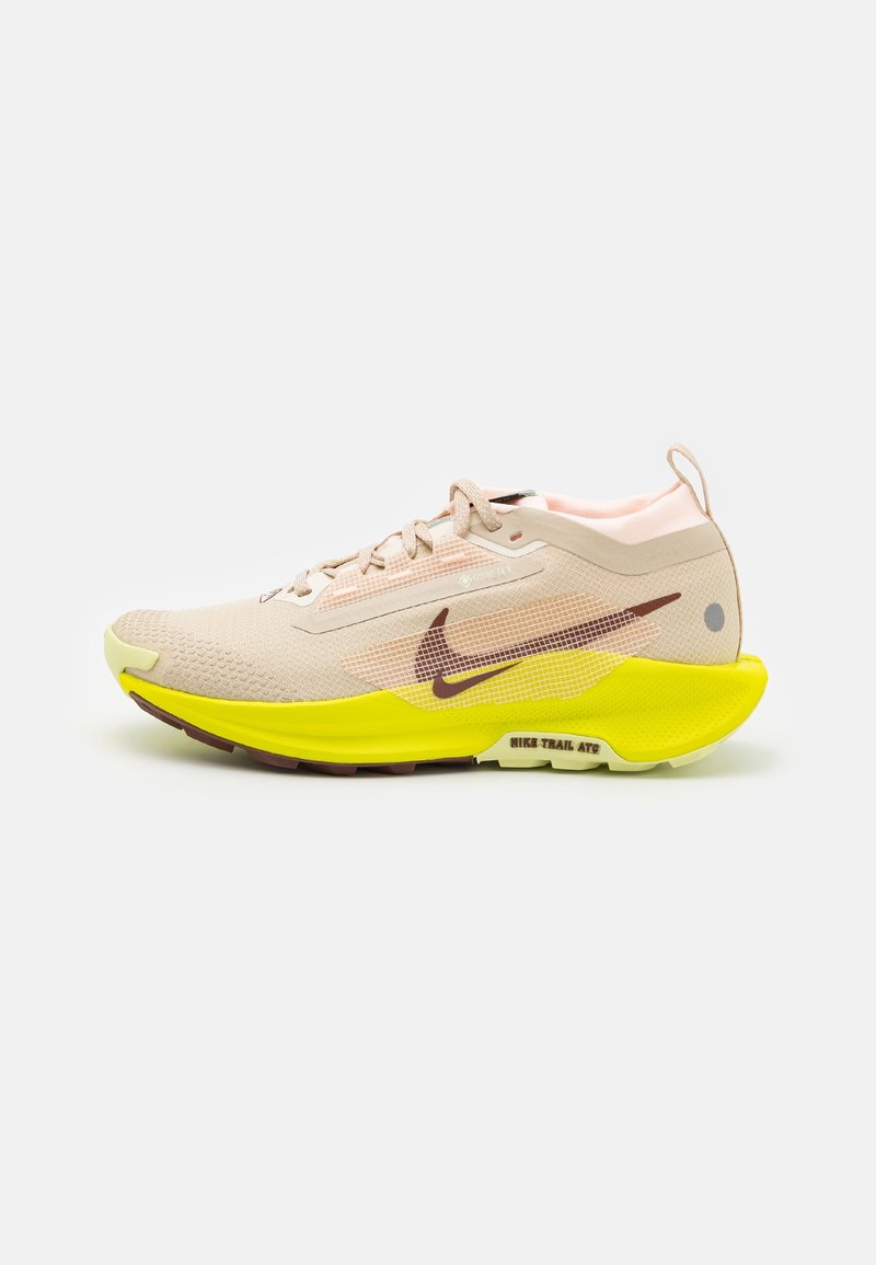 Chaussure de course Nike beige clair avec logo Swoosh marron, semelle jaune amortie et lacets sur fond blanc uni.