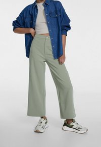 Osoba v světle zelených širokých kalhotách, bílém crop topu, oversized džínové bundě a bílých teniskách se zelenými akcenty, stojící s jednou rukou na boku.