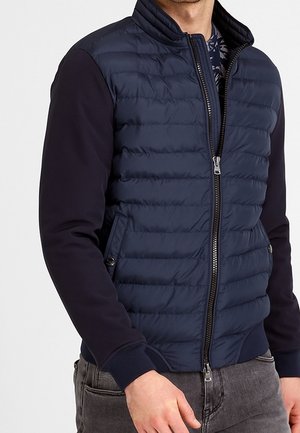 Homme portant une veste matelassée bleu marine zippée avec des manches unies foncées et un jean gris, visible de la poitrine aux hanches.