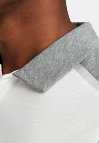 Polo bianco con colletto grigio chiaro e texture, caratterizzato da un design classico e un tessuto morbido. Primo piano dell'area del collo.