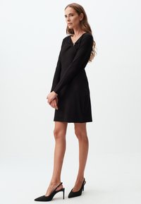 Zwarte aangesloten jurk met lange mouwen, een V-halslijn en een decoratieve strikdetail aan de voorkant, gecombineerd met zwarte slingback hakken met een puntige neus.