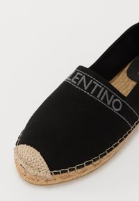 Espadrille en toile noire avec une semelle en jute texturée, présentant un embout arrondi et une bande de logo cousue le long du bord supérieur.