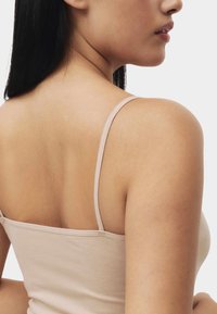 Camiseta sin mangas beige con tirantes finos, hecha de tela lisa. Presenta un diseño ajustado que enfatiza los hombros y la parte superior de la espalda.