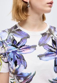 Γυναίκα με κοντά ξανθά μαλλιά που φοράει λευκό πουκάμισο με μεγάλα μοβ λουλουδένια σχέδια και μαύρα διακοσμητικά από χάντρες, καθώς και ασημένια σκουλαρίκια κρίκους.