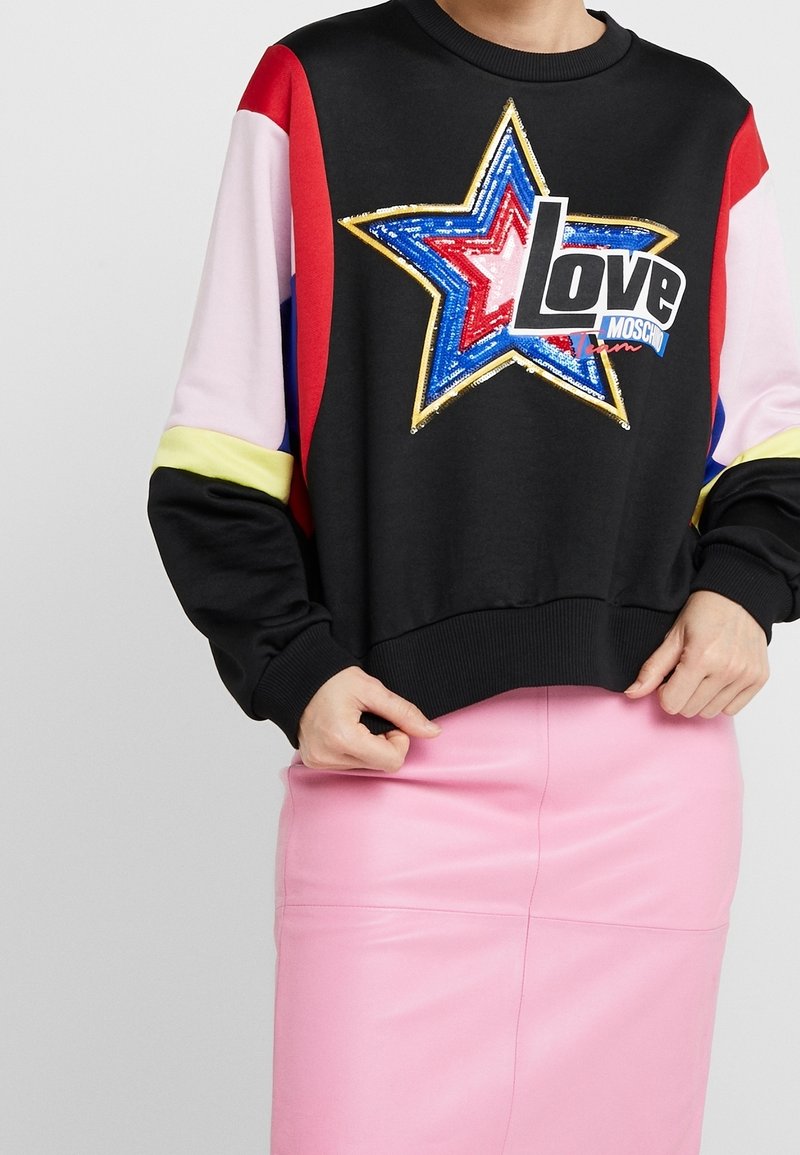 Mustat hupparit, joissa on moniväriset hihat ja iso paljettitähti muotoilu, jossa on sana "Rakkaus" paksuilla kirjaimilla. Alla vaaleanpunainen hame.