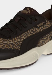 Κοντινό πλάνο ενός αθλητικού παπουτσιού Puma με μαύρα κορδόνια, leopardίσια σχέδια στα πλάγια, σκούρες λεπτομέρειες στη σόλα και μπεζ χοντρή σόλα.