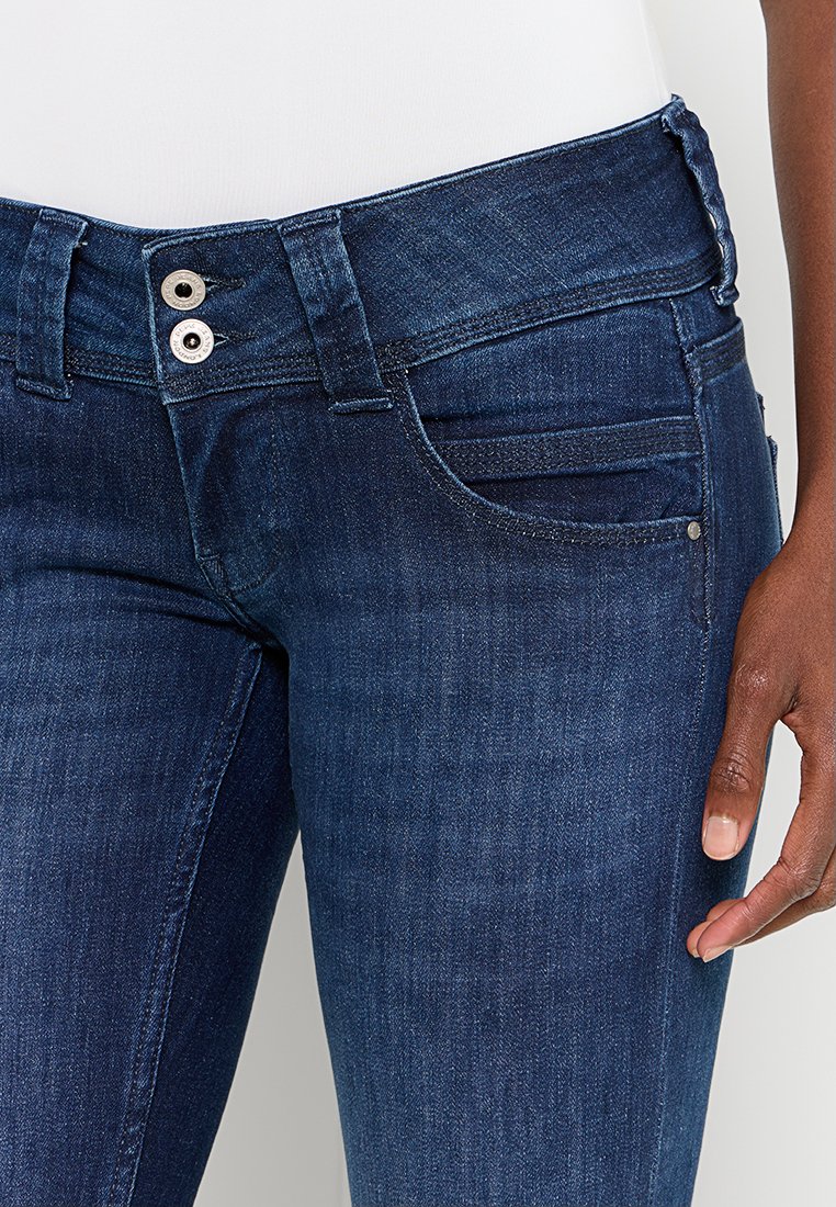 Jeans de mezclilla azul oscuro con un ajuste clásico. Presentan dos botones metálicos, bolsillos frontales y una textura suave. Diseño de pierna ajustada.