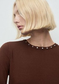 Donna bionda con un taglio di capelli bob corto guarda a sinistra, indossando un maglione marrone con piccole borchie d'argento lungo il colletto.