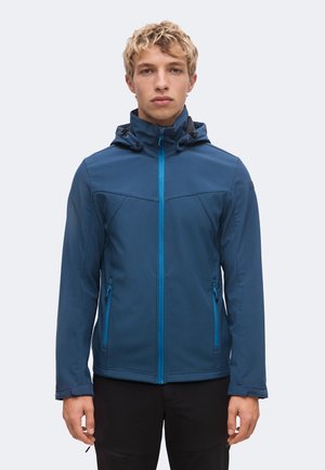 Jeune homme aux cheveux blonds courts et bouclés portant une veste à capuche bleue zippée et un pantalon noir, debout devant un fond uni.