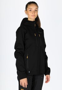 Svart softshell-jacka med huva, som har två fickor på bröstet med dragkedja och en frontdragkedja med kontrasterande guldaccenter. Slät textur.