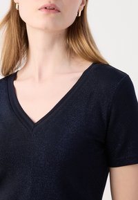Haut à col en V marine à manches courtes, fabriqué à partir d'un mat�ériau texturé qui brille subtilement. Présente un décolleté lisse et une coupe ajustée.