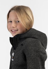 Chaqueta con capucha y cremallera en gris oscuro, hecha de una tela texturizada. Presenta un logotipo en el hombro y un detalle decorativo en la cremallera.