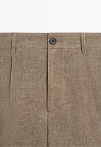 Beige Leinenhose mit schwarzem Knopfverschluss, Gürtelschlaufen und sichtbaren vorderen Falten.