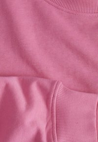 Rozā džemperis, kuram ir gluda tekstūra un ribota apkakle un piedurknes. Tuvplāns izceļ vienmērīgu šuvju un krāsas dziļumu.