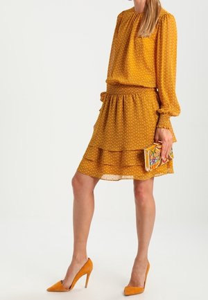 Kobieta w musztardowej sukience w groszki z długimi rękawami, trzymająca kwiatową kopertówkę i nosząca pasujące żółte szpilki.